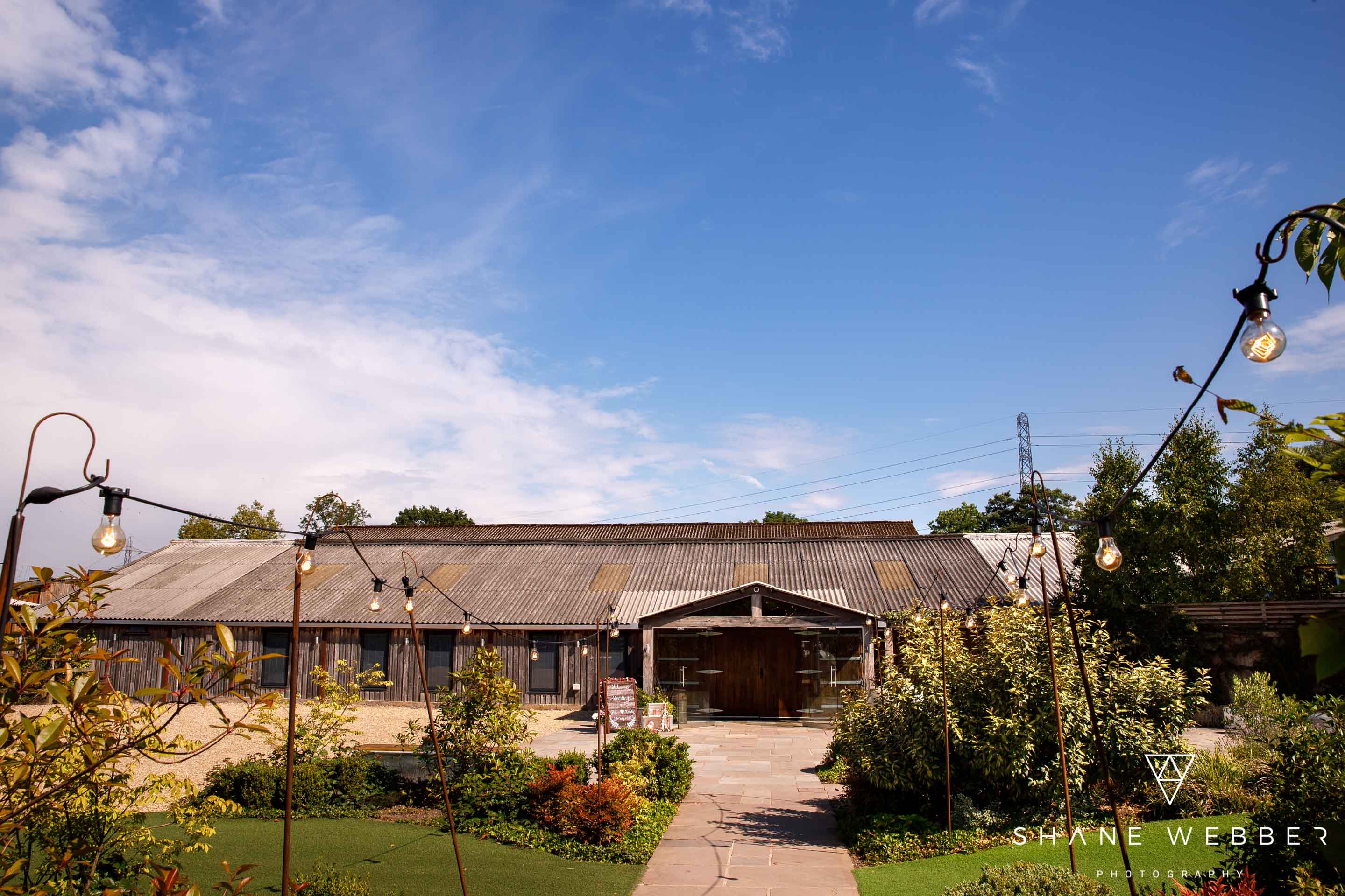 Owen House Wedding Barn from the gardens on a sunny day, festoon lights lining the path to the doors
